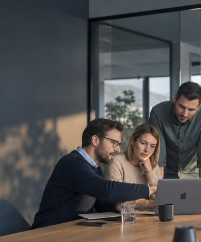 Four colleagues gathered around a laptop in a meeting room in late-afternoon light.