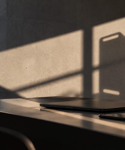 Un ordinateur portable fermé, un téléphone et des écouteurs sur un bureau sombre, dans la lumière rasante de fin de journée.