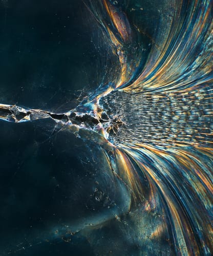 Macro d’une fissure dans une matière sombre qui se prolonge en fibres irisées, bleu et ambre.