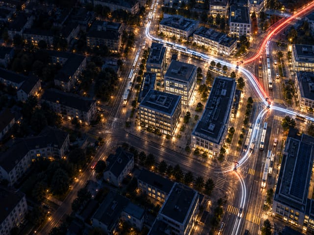 Aerial night view of an office district, a red trail of headlights veering off between the lit buildings.