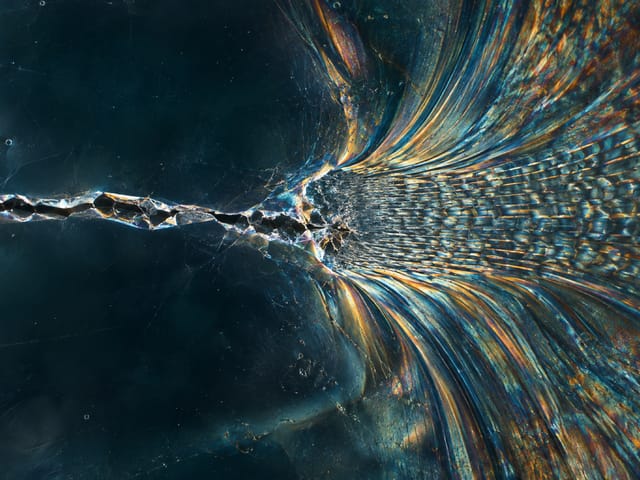 Macro of a crack in a dark material spreading into iridescent blue and amber fibres.