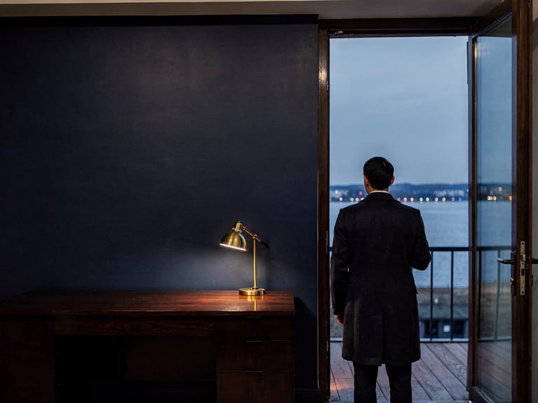 Une personne de dos à la porte ouverte d’un balcon, les lumières d’une ville au bord du lac au crépuscule.