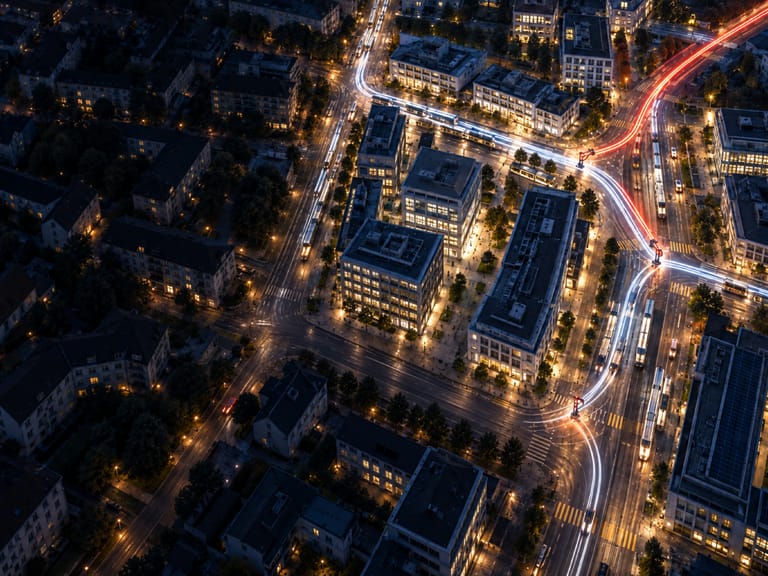 Vue aérienne nocturne d’un quartier de bureaux, une trainée rouge de phares bifurque entre les immeubles éclairés.