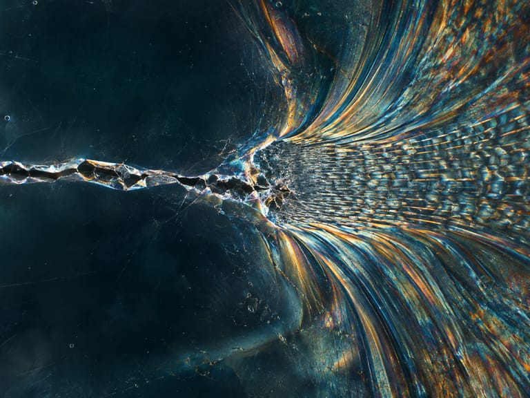 Macro d’une fissure dans une matière sombre qui se prolonge en fibres irisées, bleu et ambre.